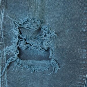 May include: Close-up of distressed denim jeans in a dark blue wash. The jeans feature a large, frayed hole, showcasing the fabric's texture and wear. The image highlights the details of the ripped design.