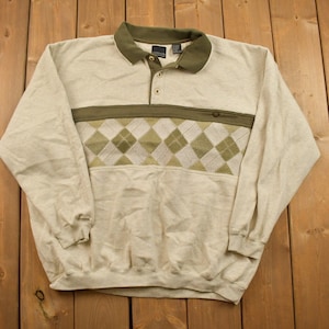 May include: A beige long-sleeved sweatshirt with a green collar and a diamond pattern on the chest. The sweatshirt has a button-up closure.
