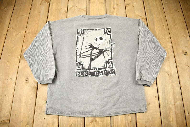 Puede incluir: Sudadera gris de manga larga con un gr&aacute;fico de Jack Skellington de The Nightmare Before Christmas, con el texto "BONE DADDY" debajo. Las mangas tienen un dise&ntilde;o texturizado y acanalado.