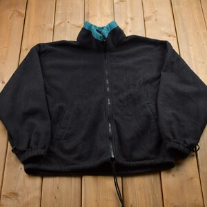 May include: A black fleece jacket with a teal zipper and collar. The jacket has a drawstring at the bottom and two pockets.