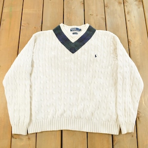 May include: Cream-colored cable knit sweater with a V-neck collar featuring a navy and green pattern. The sweater has long sleeves and a small embroidered logo on the chest. A classic, preppy style.