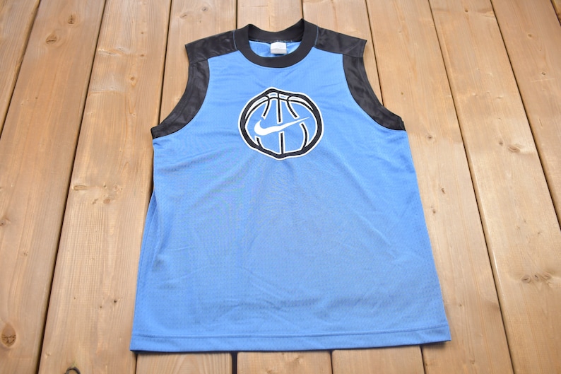 May include: A light blue sleeveless basketball jersey with black mesh side panels. The front of the jersey features a black and white basketball with a white Nike swoosh logo.