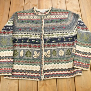 Op de afbeelding: Een grijze, groene en witte gepatroneerde cardigan met een knoopsluiting. De cardigan heeft een bloemen- en bladerenontwerp.