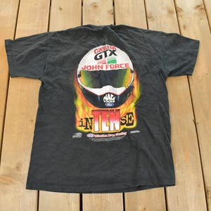 Op de afbeelding: Donkergrijs t-shirt met een racehelm-afbeelding, inclusief de tekst "Castrol GTX John Force" en "INTENSE". Het helmontwerp bevat wit, groen, geel en zwart, tegen een vurige achtergrond. Het t-shirt ligt plat.