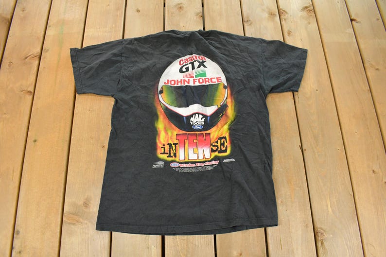 Op de afbeelding: Donkergrijs t-shirt met een racehelm-afbeelding, inclusief de tekst "Castrol GTX John Force" en "INTENSE". Het helmontwerp bevat wit, groen, geel en zwart, tegen een vurige achtergrond. Het t-shirt ligt plat.