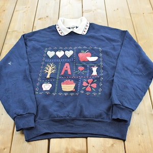 May include: A navy blue long-sleeve sweatshirt with a white collar featuring a small red heart pattern. The front displays a graphic print of apples, a basket of fruit, a tree, a flower, and the letter A, all arranged within a square border.