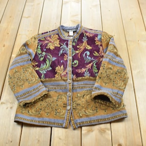 May include: A long-sleeve jacket featuring a mix of purple, gold, and light blue fabric panels with intricate floral and scrollwork patterns. The garment has a round neckline, a front button closure with light-coloured buttons, and a structured fit.