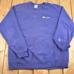 May include: A vintage Champion sweatshirt in a faded blue hue. The long-sleeved crewneck features the Champion logo embroidered in white on the chest and a small logo patch on the left sleeve cuff. The sweatshirt is laid flat on a wooden surface.