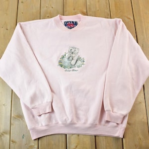 May include: A light pink sweatshirt with a crew neck and long sleeves. The front features an embroidered polar bear design with the text "Polar Bear" below it. The brand tag reads "Galt Sand".