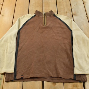 May include: A brown and tan long-sleeve pullover shirt with a quarter-zip collar. The shirt has tan sleeves and a brown body, with black trim along the seams. The collar and cuffs are ribbed.