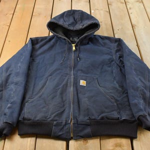 May include: A navy blue hooded jacket featuring a full-length front zip, long sleeves with ribbed cuffs, a ribbed hem, and two front pockets. A small rectangular Carhartt brand patch is stitched onto the front left pocket area.