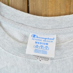Puede incluir: Etiqueta de sudadera gris Champion Reverse Weave con el texto "Champion Reverse Weave" y "Medium". La etiqueta tambi&eacute;n incluye informaci&oacute;n de talla para Canad&aacute;, M&eacute;xico, Jap&oacute;n y el pa&iacute;s de origen, El Salvador.
