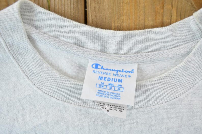 Puede incluir: Etiqueta de sudadera gris Champion Reverse Weave con el texto "Champion Reverse Weave" y "Medium". La etiqueta tambi&eacute;n incluye informaci&oacute;n de talla para Canad&aacute;, M&eacute;xico, Jap&oacute;n y el pa&iacute;s de origen, El Salvador.