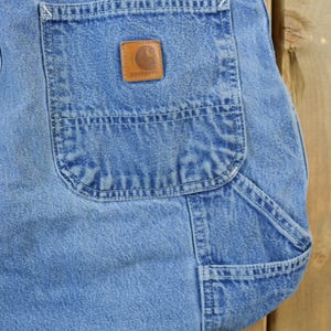 May include: A pair of blue denim carpenter jeans with a large pocket on the right leg. The pocket has a brown leather patch with the Carhartt logo.