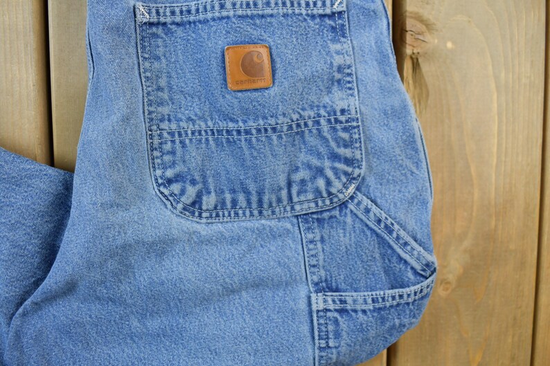 May include: A pair of blue denim carpenter jeans with a large pocket on the right leg. The pocket has a brown leather patch with the Carhartt logo.