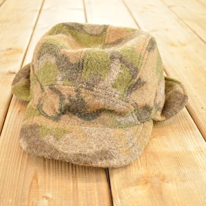 Pode incluir: Um chapéu com padrão de camuflagem com protetores de ouvido, sobre uma superfície de madeira. O chapéu apresenta uma mistura de cores verdes, castanhas e bege, com uma coroa arredondada e uma aba pequena. Os protetores de ouvido estendem-se para baixo.
