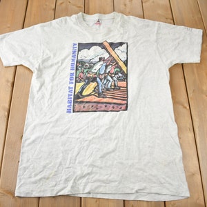 May include: A light grey t-shirt with a graphic print of people working on a house. The text "HABITAT FOR HUMANITY" is printed on the left side of the graphic.