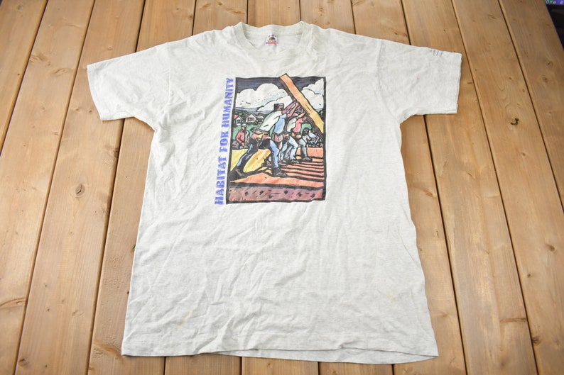 May include: A light grey t-shirt with a graphic print of people working on a house. The text "HABITAT FOR HUMANITY" is printed on the left side of the graphic.