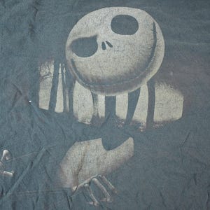 May include: Dark gray t-shirt featuring a distressed graphic of Jack Skellington from The Nightmare Before Christmas. The design includes a skeletal face, arms, and a forest silhouette. The shirt has a vintage, worn look.