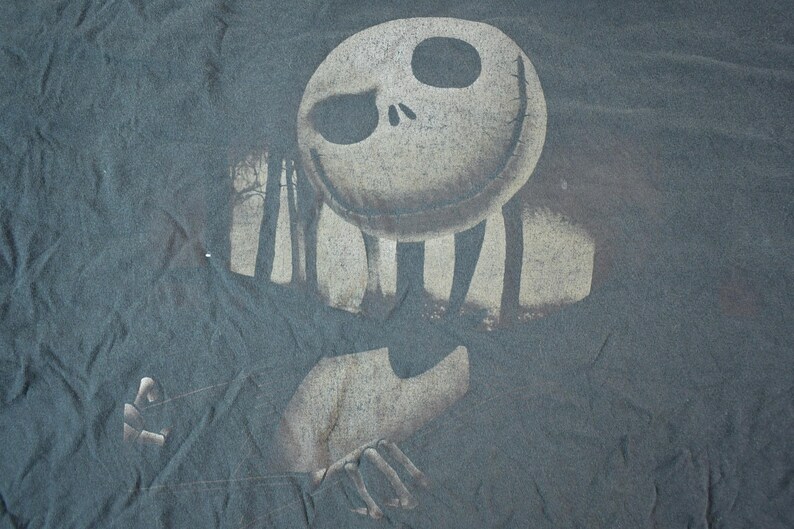 May include: Dark gray t-shirt featuring a distressed graphic of Jack Skellington from The Nightmare Before Christmas. The design includes a skeletal face, arms, and a forest silhouette. The shirt has a vintage, worn look.