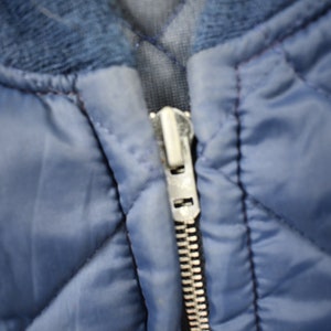 May include: A close-up of a blue quilted jacket with a silver zipper. The zipper is partially open, revealing the lining of the jacket.