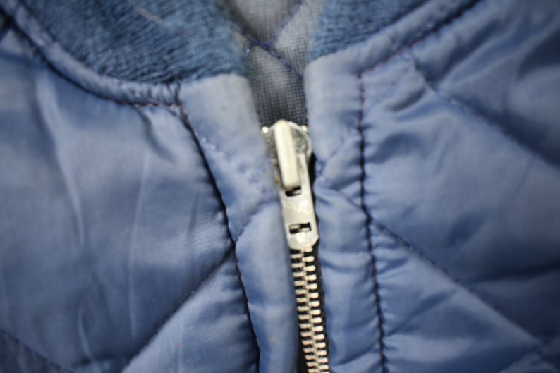 May include: A close-up of a blue quilted jacket with a silver zipper. The zipper is partially open, revealing the lining of the jacket.