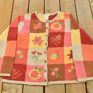 May include: A button-down cardigan sweater with a patchwork design. The sweater features embroidered floral designs in shades of red, yellow, and pink. The sweater is made of a soft material and has a cream-colored trim.