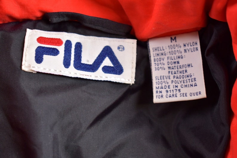 May include: A red Fila jacket with a black lining. The label says "SHELL: 100% NYLON LINING: 100% NYLON BODY FILLING: 70% DOWN 30% WATERFOWL FEATHER SLEEVE PADDING: 100% POLYESTER MADE IN CHINA RN 91175 FOR CARE SEE OVER"