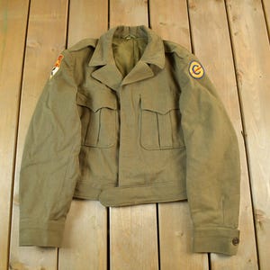 May include: Olive green military jacket with four front pockets, a notched collar, and buttoned cuffs. The jacket features patches on the sleeves, and a belt detail at the waist. The jacket is made of a durable fabric.