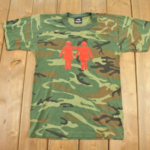 May include: A short-sleeve t-shirt featuring a green, brown, and black camouflage pattern. The center of the chest displays a graphic of two red figures standing side-by-side with one arm draped over the other's shoulder. The shirt has a crew neckline.
