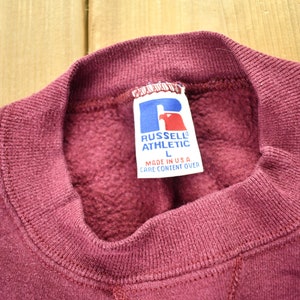 May include: Burgundy sweatshirt with a Russell Athletic logo tag. The tag is white with blue lettering and a blue logo. The tag says "Russell Athletic" and "Made in U.S.A."