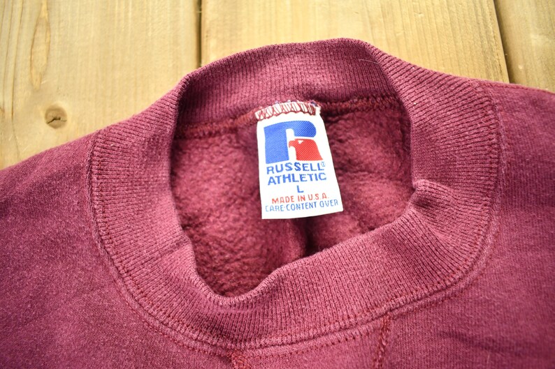 May include: Burgundy sweatshirt with a Russell Athletic logo tag. The tag is white with blue lettering and a blue logo. The tag says "Russell Athletic" and "Made in U.S.A."