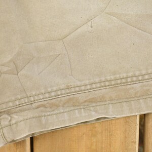 May include: Close-up of tan fabric with visible stitching and creases. The fabric appears to be part of a garment, possibly shorts or pants, with a textured surface and a worn appearance. The stitching is a contrasting color.
