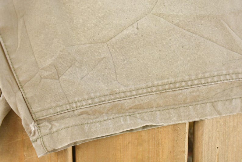 May include: Close-up of tan fabric with visible stitching and creases. The fabric appears to be part of a garment, possibly shorts or pants, with a textured surface and a worn appearance. The stitching is a contrasting color.