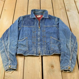 May include: A cropped blue denim jacket featuring a fold-down collar, a front zipper closure, and two front pockets. The jacket has a red and white checkered lining visible at the neckline and long sleeves with metal button cuffs at the wrists.