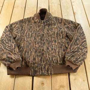 May include: A brown and black animal print bomber jacket featuring a textured fabric, a ribbed brown collar, ribbed brown cuffs, and a ribbed brown hem. The jacket has a front zipper closure and long sleeves, showcasing a consistent pattern throughout.
