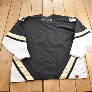 Puede incluir: Camiseta de hockey negra con detalles en blanco y dorado. La camiseta tiene un logotipo en la parte delantera que dice "KOHO".