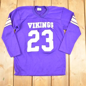 May include: A purple long-sleeved jersey with the word "VIKINGS" and the number "23" in white. The sleeves have white and tan stripes. The jersey is made of a soft material and has a V-neck.