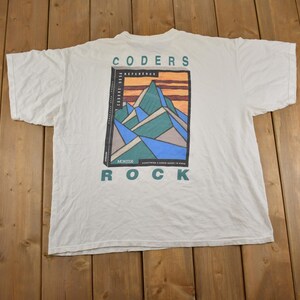 Puede incluir: Camiseta blanca con un gráfico de montañas y el texto "CODERS ROCK" impreso en la espalda.