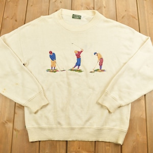 Può includere: Una felpa color crema con collo rotondo e maniche lunghe. La felpa presenta un disegno ricamato di tre golfisti in diverse pose.