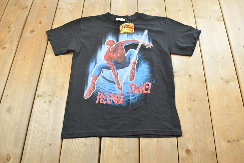 May include: Black t-shirt with a Spider-Man graphic, showing the superhero in a dynamic pose with the words "HANG TIME!" in red. The character is in his red and blue suit against a blue backdrop. A tag is attached to the shirt.