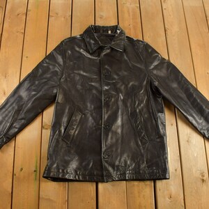 May include: A black leather jacket with a button closure and two pockets. The jacket is laid flat on a wooden surface.