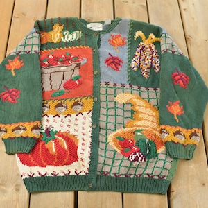 May include: Green cardigan with a patchwork design featuring autumn harvest motifs. The sweater includes images of pumpkins, leaves, and a basket of apples. The sweater has a button-up front and long sleeves.