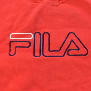 Puede incluir: Primer plano de una sudadera roja con el nombre de la marca "FILA" bordado en blanco y azul marino. Las letras est&aacute;n delineadas con hilo azul marino, creando un logotipo llamativo. La tela parece suave y c&oacute;moda.