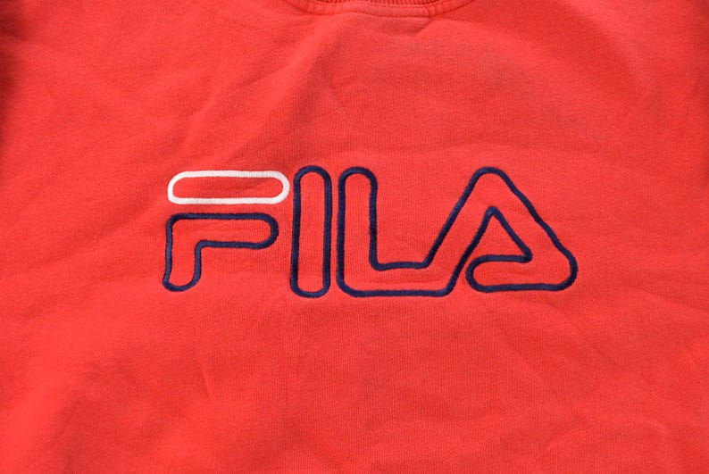 Puede incluir: Primer plano de una sudadera roja con el nombre de la marca "FILA" bordado en blanco y azul marino. Las letras est&aacute;n delineadas con hilo azul marino, creando un logotipo llamativo. La tela parece suave y c&oacute;moda.
