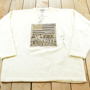 May include: Cream-colored long-sleeve sweatshirt featuring a graphic of an American flag and the text "Avirex Tuskegee Airmen." The sweatshirt has a crew neck and a tag attached near the collar.