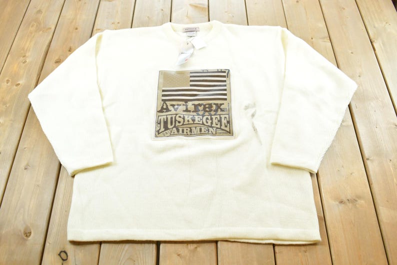 May include: Cream-colored long-sleeve sweatshirt featuring a graphic of an American flag and the text "Avirex Tuskegee Airmen." The sweatshirt has a crew neck and a tag attached near the collar.