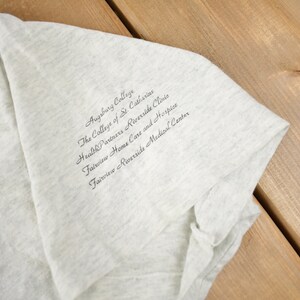 May include: A grey t-shirt with the text "Augsburg College, The College of St. Catherine, Health Partners Riverside Clinic, Fairview Home Care and Hospice, Fairview Riverside Medical Center" printed on the front.