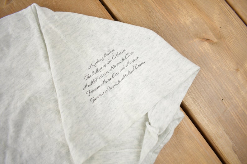 May include: A grey t-shirt with the text "Augsburg College, The College of St. Catherine, Health Partners Riverside Clinic, Fairview Home Care and Hospice, Fairview Riverside Medical Center" printed on the front.