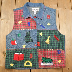 May include: A blue denim vest with a quilted design and patchwork squares. The squares feature a red and green plaid pattern, a blue denim pattern, and a red plaid pattern. The vest has a collar and button closure. The vest is decorated with embroidered images of a cat, an apple, a book, a pencil, a bell, a ruler, and the letters A, B, C, D, 1, 2, 3, and 4.